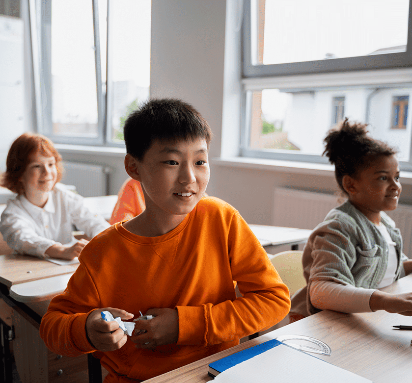 a boy sitting at a desk in a classroom full of children